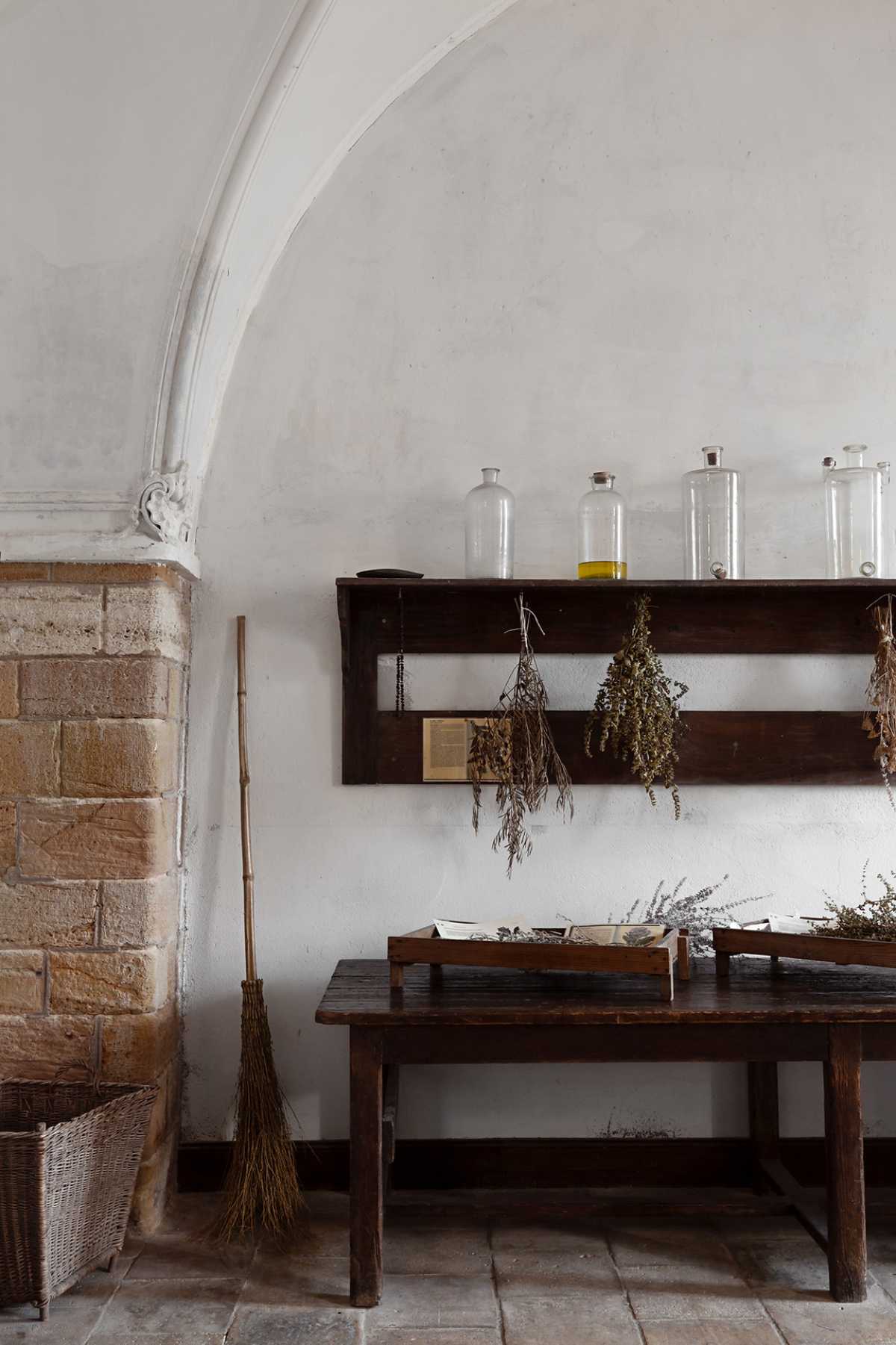 Detalle de parte de una cocina con mesa de madera oscura y estantería con botes de cristal encima y ramas de planta secando