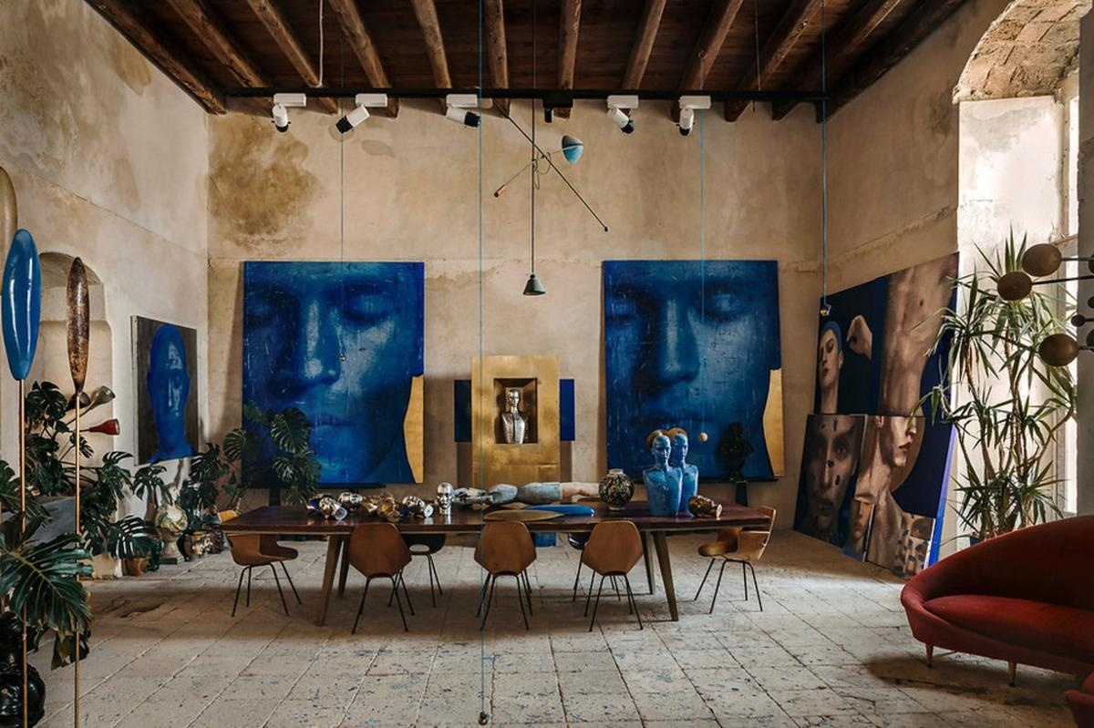 Estudio de un artista lleno de cuadros apoyados o colgados en pared en colores azules y mesa de madera amplia situada en el centro con sillas doradas. Sobre la mesa hay figuras y elementos del artista.