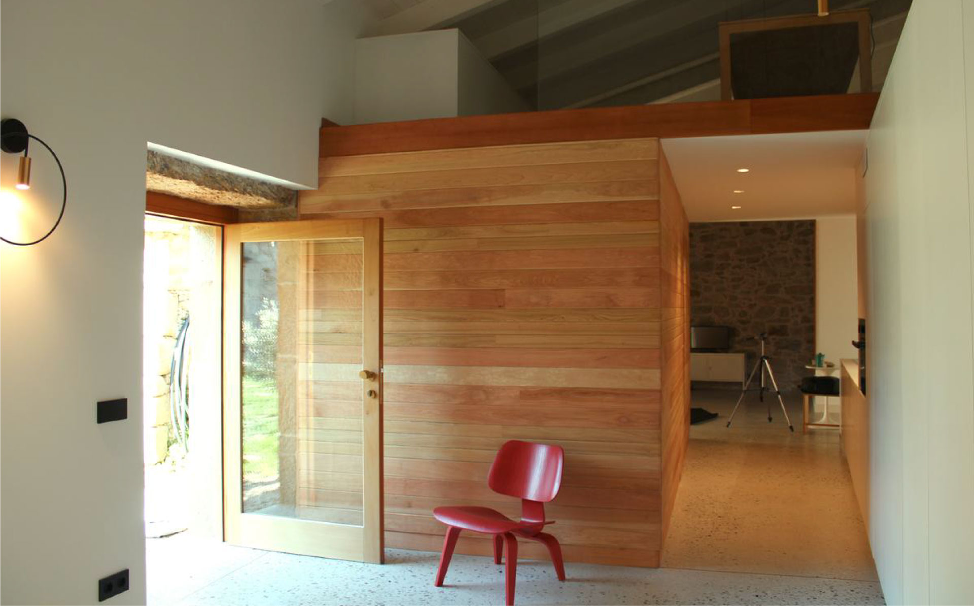 Interior de dos plantas, paredes blancas y de madera, puerta de cristal y silla roja.