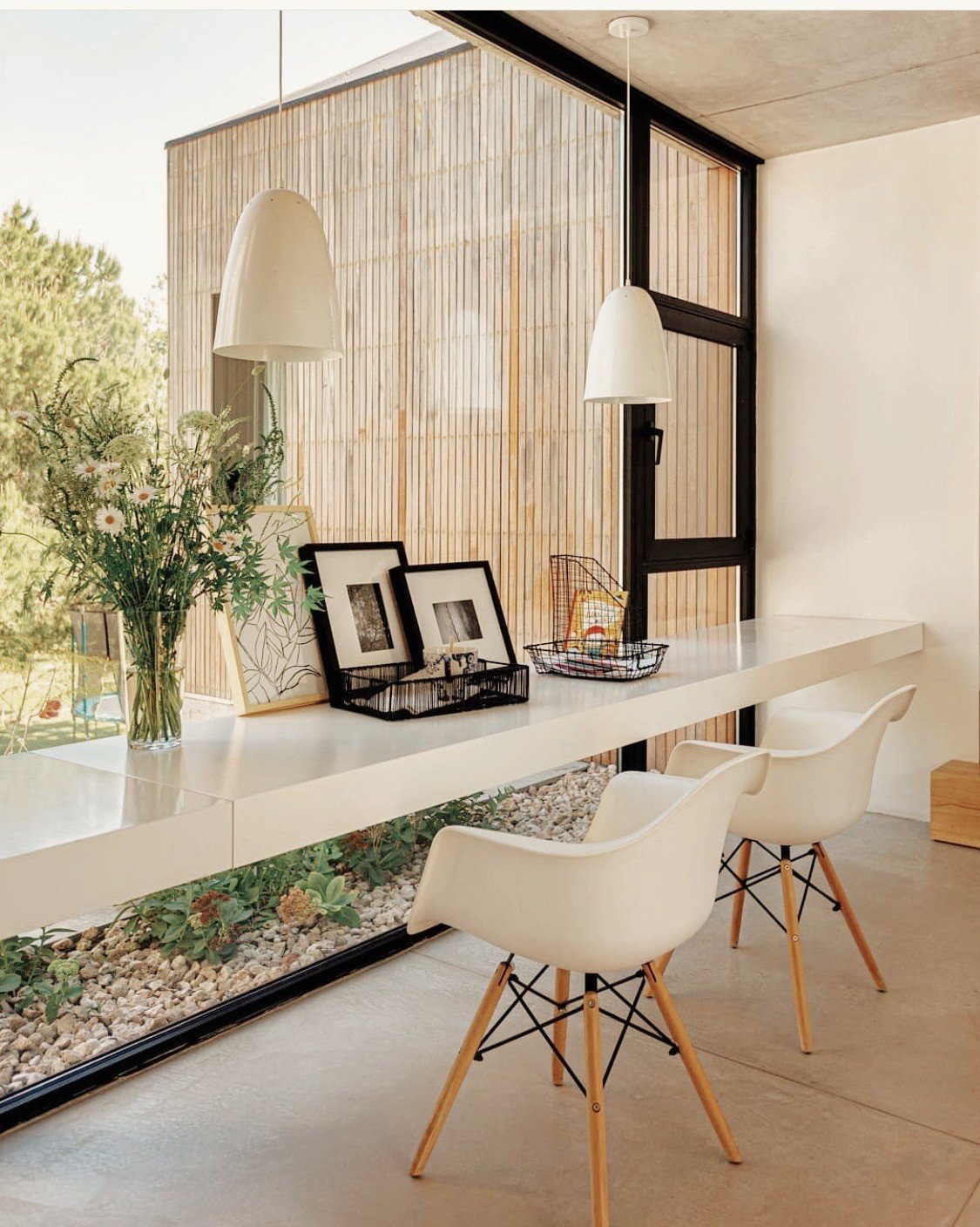 Espacio de home office con escritorio flotante blanco frente a una ventana panorámica. Decoración con flores, marcos, cesta metálica y lámparas colgantes. Dos sillas blancas con patas de madera. Vista exterior de jardín con piedras y vegetación